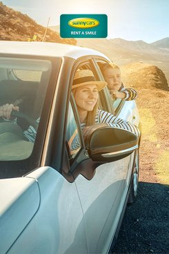 Mutter und Sohn sitzen in einem weißen Auto mit Blick auf das wunderschöne Meer. Verlinkung zu SunnyCars Angebote mit 15 Euro Rabatt.